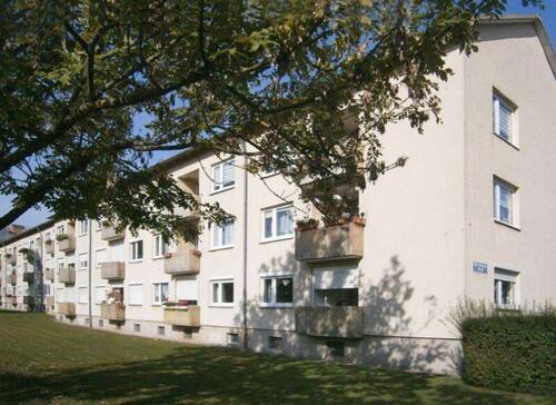 Rückansicht des mehrstöckigen Wohngebäudes mit Balkonen, umgeben von Grünanlagen mit Bäumen und grünen Sträuchern - Nachmieterin für SINGLEWOHNUNG in ruhiger Lage gesucht!