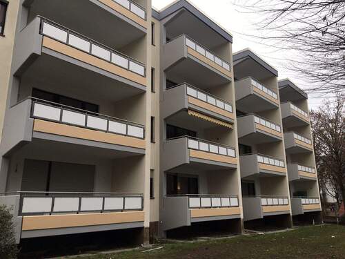 Frontansicht eines mehrstöckigen Gebäudes mit Balkonen, umgeben von Wiesen und Bäumen. - 