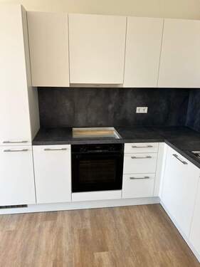 Beispiel einer L-förmigen Küche mit weißen Unterschränken und grifflose, helle Oberschränke. Die Arbeitsplatte sowie der Spritzschutz an der Wand sind aus einem anthrazitfarbenen Material. Der Boden ist ein Vinylboden in Holzoptik. - 