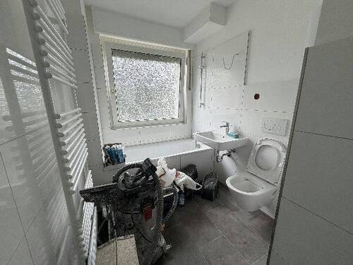 Blick in Bad mit weißen Wandfliesen, weißem Waschbecken, grauen Bodenfliesen, Hänge-WC und Badewanne mit Duschvorrichtung und Fenster - 3 Zimmer Etagenwohnung in Meerbusch