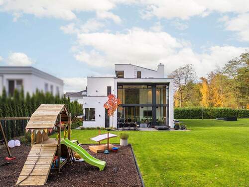 Modernes, weißes Haus mit großer Glasfront und Terrasse, davor ein Garten mit Spielplatz, Rasen und herbstlichen Bäumen im Hintergrund - 