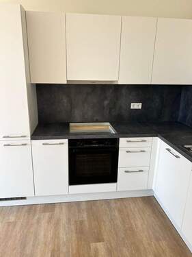 Beispiel einer L-förmigen Küche mit weißen Unterschränken und grifflose, helle Oberschränke. Die Arbeitsplatte sowie der Spritzschutz an der Wand sind aus einem anthrazitfarbenen Material. Der Boden ist ein Vinylboden in Holzoptik. - 