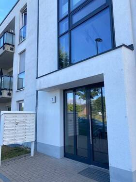 Blick auf den Hauseingang des weißen Hauses mit grauer Glaseingangstür, Balkonen und grauen Fenstern - 160047215198 C4 'Helle 2-Zimmer-Wohnung in Meerbusch-Osterath - im EG mit Garten'