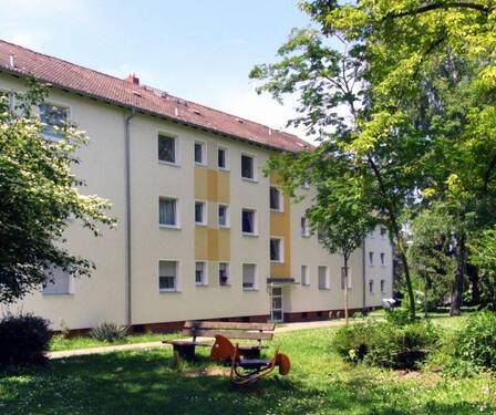 Hausansicht eines mehrstöckigen Wohngebäudes umgeben von Wiese, Sträuchern und Bäumen. - Nachmieter für 2-Zimmer-Wohnung gesucht!