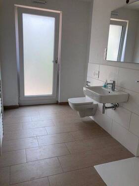 Tageslicht Duschbadezimmer mit weißem wandhängendem WC, Waschbecken. Die Wände sind im Nassbereich weiß gefliest. Der Boden ist hell gefliest. - 