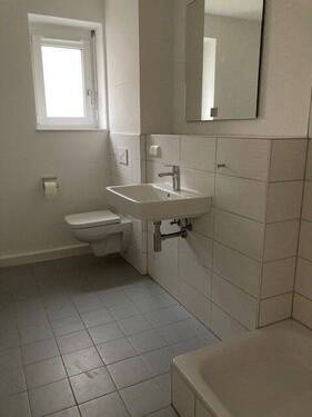 Tageslicht Duschbadezimmer mit weißem wandhängendem WC, Waschbecken. Die Wände sind im Nassbereich wandhoch und weiß gefliest. Der Boden ist hellgrau gefliest. - 