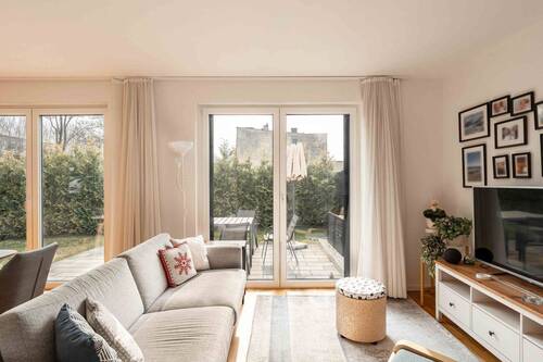 Heller Wohnbereich mit einem Fernseher an der rechten Wand, einer grauen Couch gegenüber und Zugang zur Terrasse - 5 Zimmer Doppelhaushälfte in Zossen-Wünsdorf