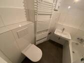 Blick in Bad mit weißen Wandfliesen, weißem Waschbecken, grauen Bodenfliesen, Hänge-WC und Badewanne mit Duschvorrichtung - 