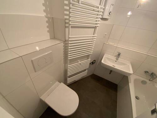 Blick in Bad mit weißen Wandfliesen, weißem Waschbecken, grauen Bodenfliesen, Hänge-WC und Badewanne mit Duschvorrichtung - 