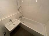 Blick in Bad mit weißen Wandfliesen, weißem Waschbecken, grauen Bodenfliesen, Hänge-WC und Badewanne mit Duschvorrichtung - 