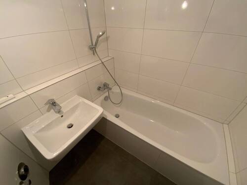 Blick in Bad mit weißen Wandfliesen, weißem Waschbecken, grauen Bodenfliesen, Hänge-WC und Badewanne mit Duschvorrichtung - 