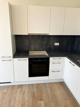 Beispiel einer L-förmigen Küche mit weißen Unterschränken und grifflose, helle Oberschränke. Die Arbeitsplatte sowie der Spritzschutz an der Wand sind aus einem anthrazitfarbenen Material. Der Boden ist ein Vinylboden in Holzoptik. - 