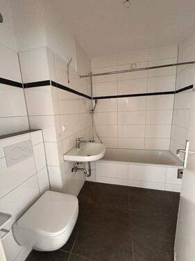 Badezimmer mit grauen Bodenfliesen, weißen Wandfliesen mit Schwarzer Bodüre, sowie weißem Waschbecken, Hänge WC und Badewanne.jpg - 