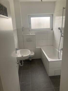 Tageslicht Badezimmer mit Badewanne und Duschvorrichtung. Weißes wandhängendes WC und Waschbecken. Die Wände sind im Nassbereich raumhoch weiß gefliest. Der Boden ist dunkel gefliest. - 3 Zimmer Etagenwohnung in Sulzbach