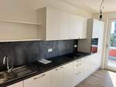 Beispiel einer Küche mit weißen Unterschränken und grifflose, helle Oberschränke. Arbeitsplatte und Spritzschutz sind in einem anthrazitfarbenen Material gehalten. Mittig ist ein Kochfeld eingebaut, rechts ein hoher Schrank mit integrieten Backofen. - Etagenwohnung mit 120,50 m&sup2; in Frankfurt am Main zur Miete