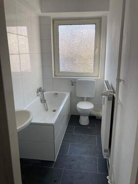 Bad mit Badewanne, weißen Sanitärobjekten, weißem Fliesenspiegel und dunkelgrauen Bodenfliesen - 