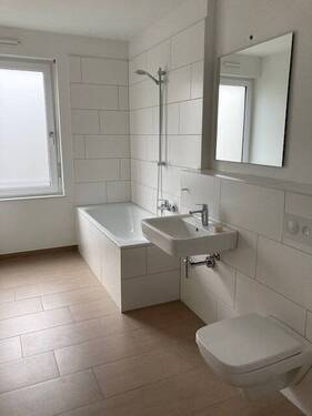 Tageslicht Badezimmer mit Dusche, weißem wandhängendem WC, Waschbecken und Handtuch Heizkörper. Die Wände sind weiß gefliest. Der Boden ist dunkel gefliest. - Beispielfoto - 