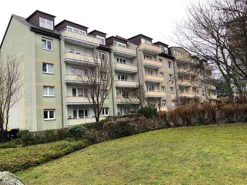 Rückansicht des mehrstöckigen Wohngebäudes mit Balkonen, umgeben von Grünanlagen mit Bäumen und grünen Sträuchern - 