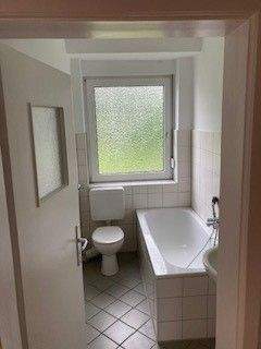 Blick in Bad mit weißen Wandfliesen, weißem Waschbecken, grauen Bodenfliesen, Hänge-WC und Badewanne mit Duschvorrichtung und Fenster - 