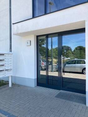 Blick auf den Hauseingang des weißen Hauses mit grauer Glaseingangstür - 16004721567 C25 'Wohnen über den Dächern: Stilvolle 3-Zimmer-Staffelgeschosswohnung'