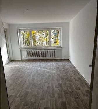 Wohnzimmer mit großer Fensterfront in die Grünanlage, Zugang zum Balkon, helle Wände und heller Bodenbelag in Holzoptik.JPG - 