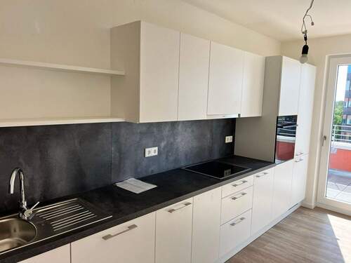 Beispiel einer Küche mit weißen Unterschränken und grifflose, helle Oberschränke. Arbeitsplatte und Spritzschutz sind in einem anthrazitfarbenen Material gehalten. Mittig ist ein Kochfeld eingebaut, rechts ein hoher Schrank mit integrieten Backofen. - 