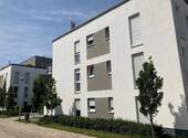 Seitenansicht eines modernen, mehrstöckigen Wohngebäudes mit heller Putzfassade und grauen Akzentflächen, davor begrünte Außenanlage mit Bäumen, Sträuchern und gepflastertem Gehweg - 2 Zimmer Etagenwohnung in Kelsterbach