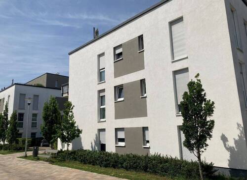 Seitenansicht eines modernen, mehrstöckigen Wohngebäudes mit heller Putzfassade und grauen Akzentflächen, davor begrünte Außenanlage mit Bäumen, Sträuchern und gepflastertem Gehweg - 2 Zimmer Etagenwohnung in Kelsterbach