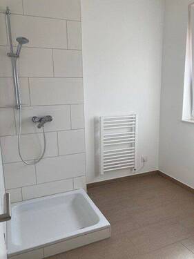 Tageslicht Bad mit Dusche mit weißem wandhängendem WC, Waschbecken und Handtuch Heizkörper. Die Wände sind im Nassbereich raumhoch und weiß gefliest. Der Boden ist hell gefliest. - 