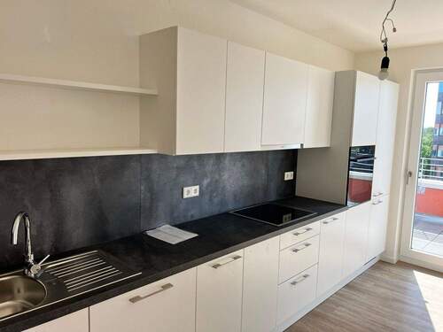 Beispiel einer Küche mit weißen Unterschränken und grifflose, helle Oberschränke. Arbeitsplatte und Spritzschutz sind in einem anthrazitfarbenen Material gehalten. Mittig ist ein Kochfeld eingebaut, rechts ein hoher Schrank mit integrieten Backofen. - 