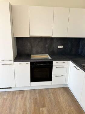 Beispiel einer L-förmigen Küche mit weißen Unterschränken und grifflose, helle Oberschränke. Die Arbeitsplatte sowie der Spritzschutz an der Wand sind aus einem anthrazitfarbenen Material. Der Boden ist ein Vinylboden in Holzoptik. - 