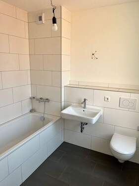 Beispiel eines Badezimmers mit weißem Wand-WC, weißem Wandwaschbecken und Badewanne mit weißen Wandfliesen auf anthrazitfarbenen Fliesenboden - 