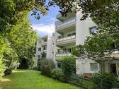 Rückansicht eines mehrstöckigen Wohngebäudes mit Gärten und Balkonen, umgeben von Bäumen und Sträuchern. - Schöne 1,5-Zimmer-Wohnung mit Balkon zu vermieten!