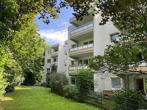 Rückansicht eines mehrstöckigen Wohngebäudes mit Gärten und Balkonen, umgeben von Bäumen und Sträuchern. - Schöne 1,5-Zimmer-Wohnung mit Balkon zu vermieten!