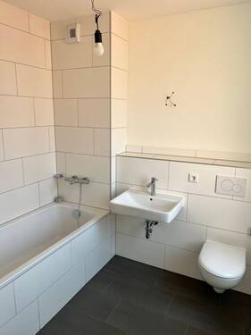 Beispiel eines Badezimmers mit weißem Wand-WC, weißem Wandwaschbecken und Badewanne mit weißen Wandfliesen auf anthrazitfarbenen Fliesenboden - 