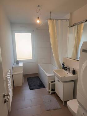 Tageslicht Wannen Badezimmer mit weißem wandhängenden WC, Waschbecken und Handtuchheizkörper. Die Wände sind im Nassbereich raumhoch und weiß gefliest. Der Boden ist hell gefliest. - 