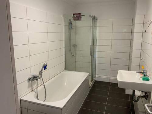 Badezimmer mit weißen Fliesen an den Wänden und dunklen Fußbodenfliesen. Badewanne und Dusche hintereinander eingebaut. Rechte Seite befindet sich das Waschbecken. - 